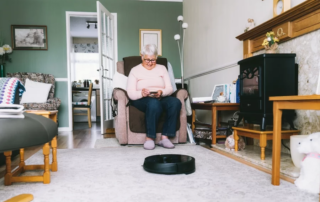 Senior woman using a robot vacuum cleaner through phone settings while sitting on armchair